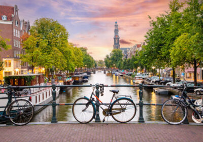 bridge-over-an-amsterdam-canal-in-sunset-1 Sunset in Amsterdam - Bicycles parked on a bridge over a canal in the city centre, with typical Dutch houses on the background. Streets illuminated with lights.